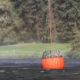 Large orange circular buoy floating on calm water with ropes extending upward, forested shore in background.