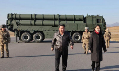 Man in a brown leather jacket and a woman in a black coat walk on a tarmac in front of a large green missile launcher truck with soldiers nearby.