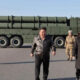Man in a brown leather jacket and a woman in a black coat walk on a tarmac in front of a large green missile launcher truck with soldiers nearby.
