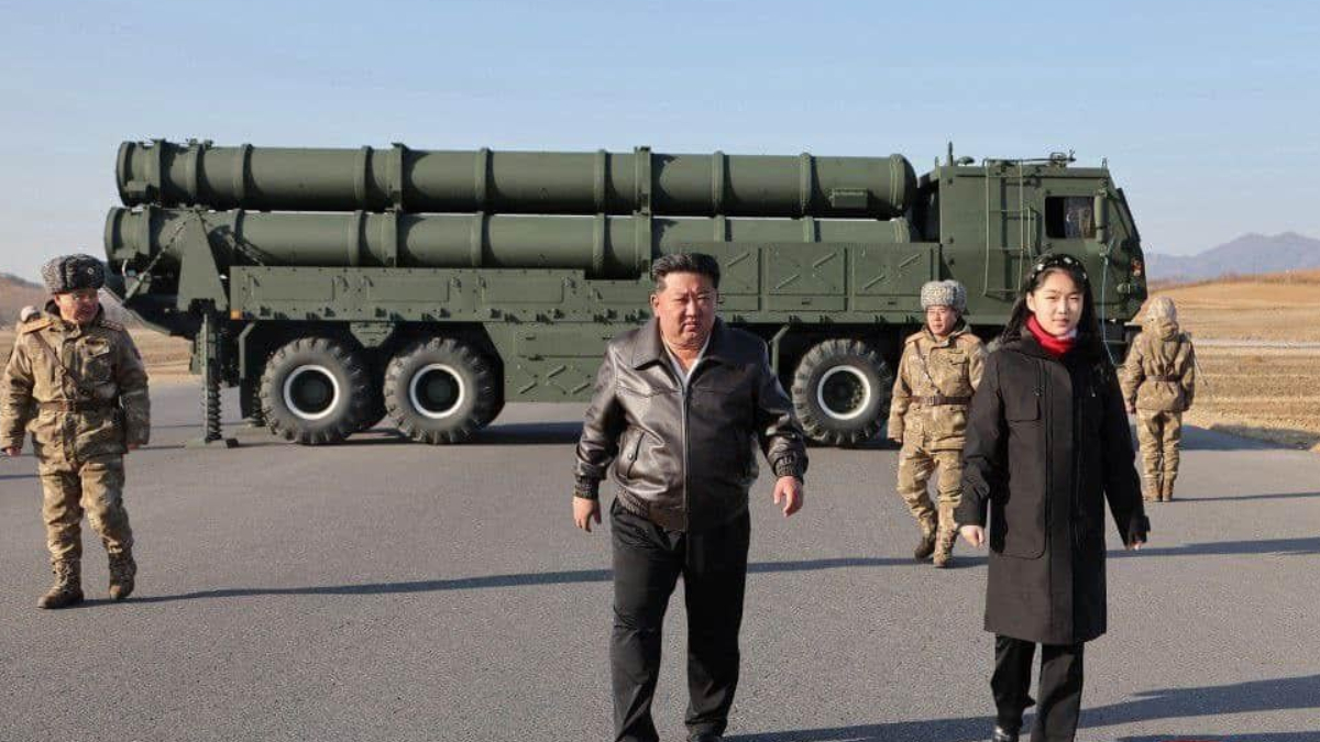 Man in a brown leather jacket and a woman in a black coat walk on a tarmac in front of a large green missile launcher truck with soldiers nearby.