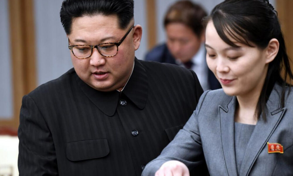Two officials in a formal setting look at documents together; man in a dark pinstripe suit with glasses on the left, woman in a gray suit with a red lapel badge on the right.