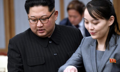 Two officials in a formal setting look at documents together; man in a dark pinstripe suit with glasses on the left, woman in a gray suit with a red lapel badge on the right.