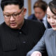 Two officials in a formal setting look at documents together; man in a dark pinstripe suit with glasses on the left, woman in a gray suit with a red lapel badge on the right.