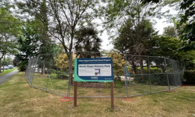Chain-link fence surrounds a small park area with a sign about the North Slope Historic Park project in the foreground.