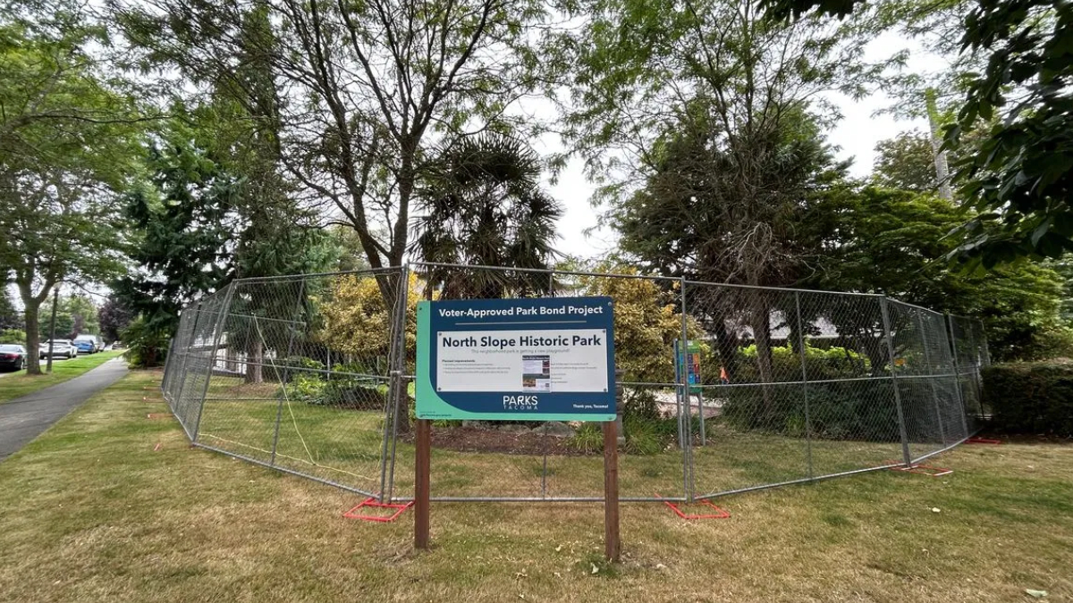 Chain-link fence surrounds a small park area with a sign about the North Slope Historic Park project in the foreground.