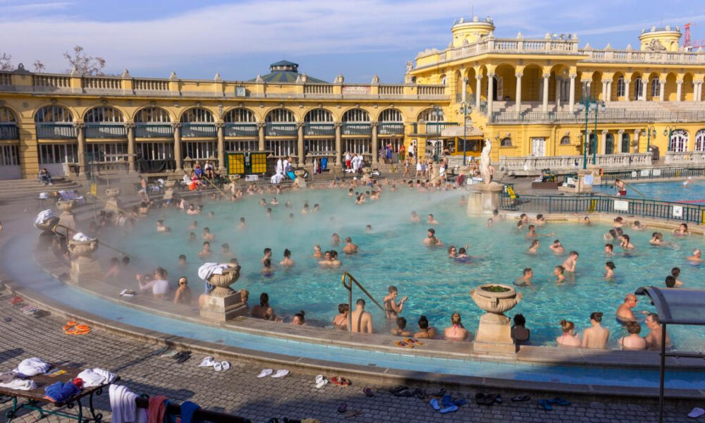 Crowded outdoor thermal pool with many swimmers amid a yellow historic bathhouse facade.