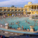 Crowded outdoor thermal pool with many swimmers amid a yellow historic bathhouse facade.
