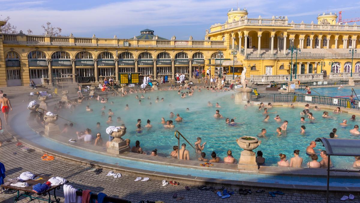 Crowded outdoor thermal pool with many swimmers amid a yellow historic bathhouse facade.