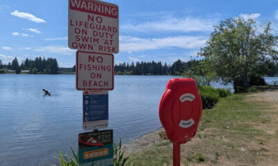 Lakeside scene with large warning signs about no lifeguard, swim at your own risk, and no fishing on the beach; a red life-rescue cabinet nearby.