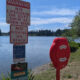 Lakeside scene with large warning signs about no lifeguard, swim at your own risk, and no fishing on the beach; a red life-rescue cabinet nearby.