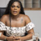 Woman with braids wearing an off‑the‑shoulder floral dress sits at a desk, hands folded, speaking to someone off‑frame.