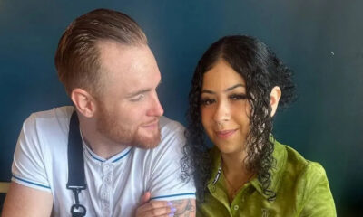 Two people sit close together, smiling at each other; a bearded man in a white shirt with an apron and a woman with curly dark hair in a green blouse sit against a dark background.