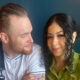 Two people sit close together, smiling at each other; a bearded man in a white shirt with an apron and a woman with curly dark hair in a green blouse sit against a dark background.