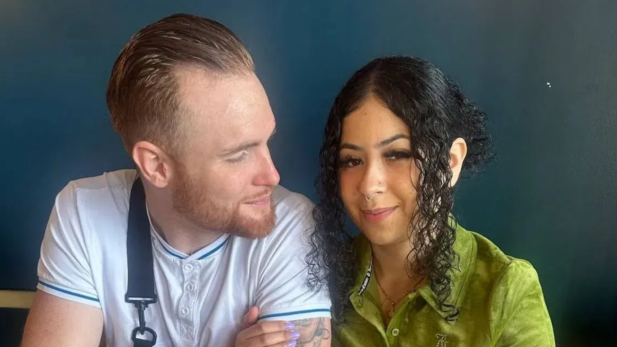 Two people sit close together, smiling at each other; a bearded man in a white shirt with an apron and a woman with curly dark hair in a green blouse sit against a dark background.