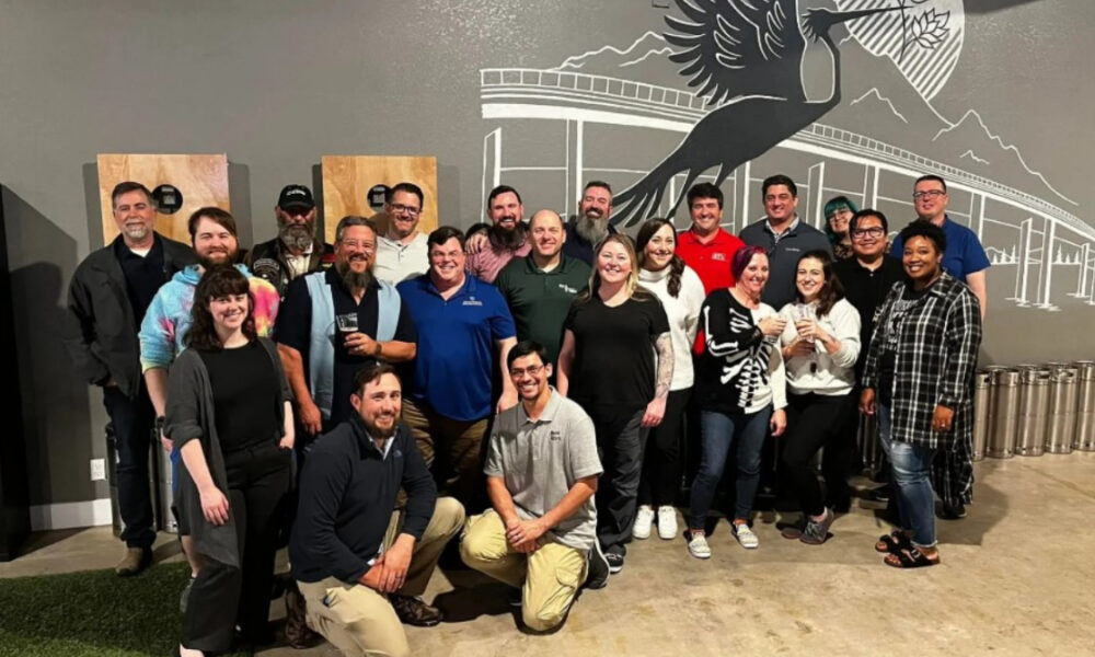 Group of coworkers posing for a photo in a brewery/event space, with a large bird mural on the wall behind them.