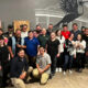 Group of coworkers posing for a photo in a brewery/event space, with a large bird mural on the wall behind them.
