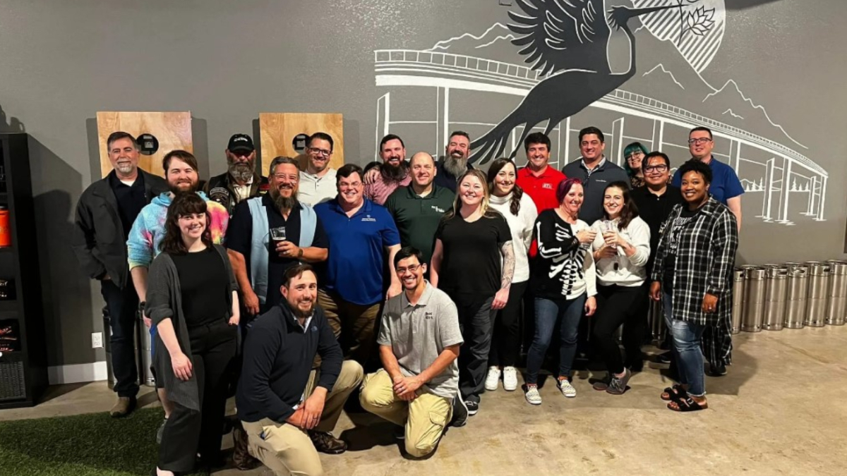 Group of coworkers posing for a photo in a brewery/event space, with a large bird mural on the wall behind them.
