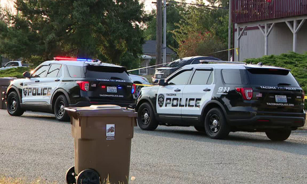 Two Tacoma Police Department SUVs with red and blue lights flashing, parked on a residential street with a trash bin in the foreground.