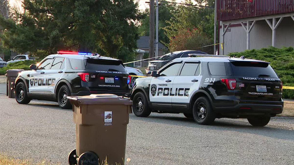 Two Tacoma Police Department SUVs with red and blue lights flashing, parked on a residential street with a trash bin in the foreground.