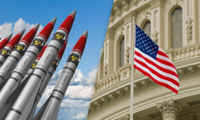 Row of missiles pointed upward beside a waving U.S. flag in front of a grand government building.