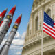 Row of missiles pointed upward beside a waving U.S. flag in front of a grand government building.