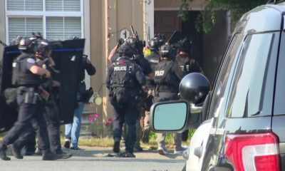 Heavily armed police officers in tactical gear and helmets move toward a house entrance, shields raised, as a police SUV sits parked in the foreground.