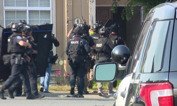Heavily armed police officers in tactical gear and helmets move toward a house entrance, shields raised, as a police SUV sits parked in the foreground.