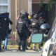 Heavily armed police officers in tactical gear and helmets move toward a house entrance, shields raised, as a police SUV sits parked in the foreground.