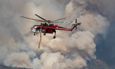 Red firefighting helicopter hovering amid thick smoke, releasing a long orange water hose toward a wildfire.