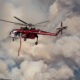 Red firefighting helicopter hovering amid thick smoke, releasing a long orange water hose toward a wildfire.