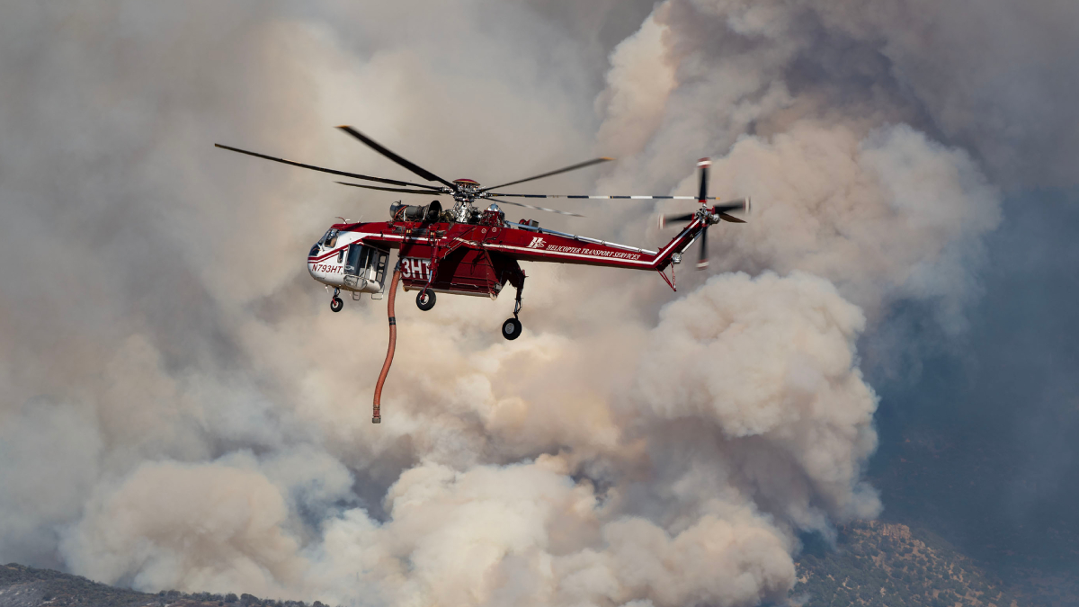 Red firefighting helicopter hovering amid thick smoke, releasing a long orange water hose toward a wildfire.