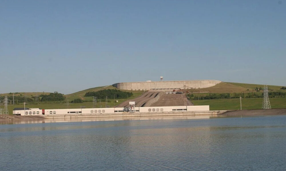 Hydroelectric facility by a lake with a long white building and grassy hill behind a concrete dam structure.