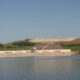 Hydroelectric facility by a lake with a long white building and grassy hill behind a concrete dam structure.