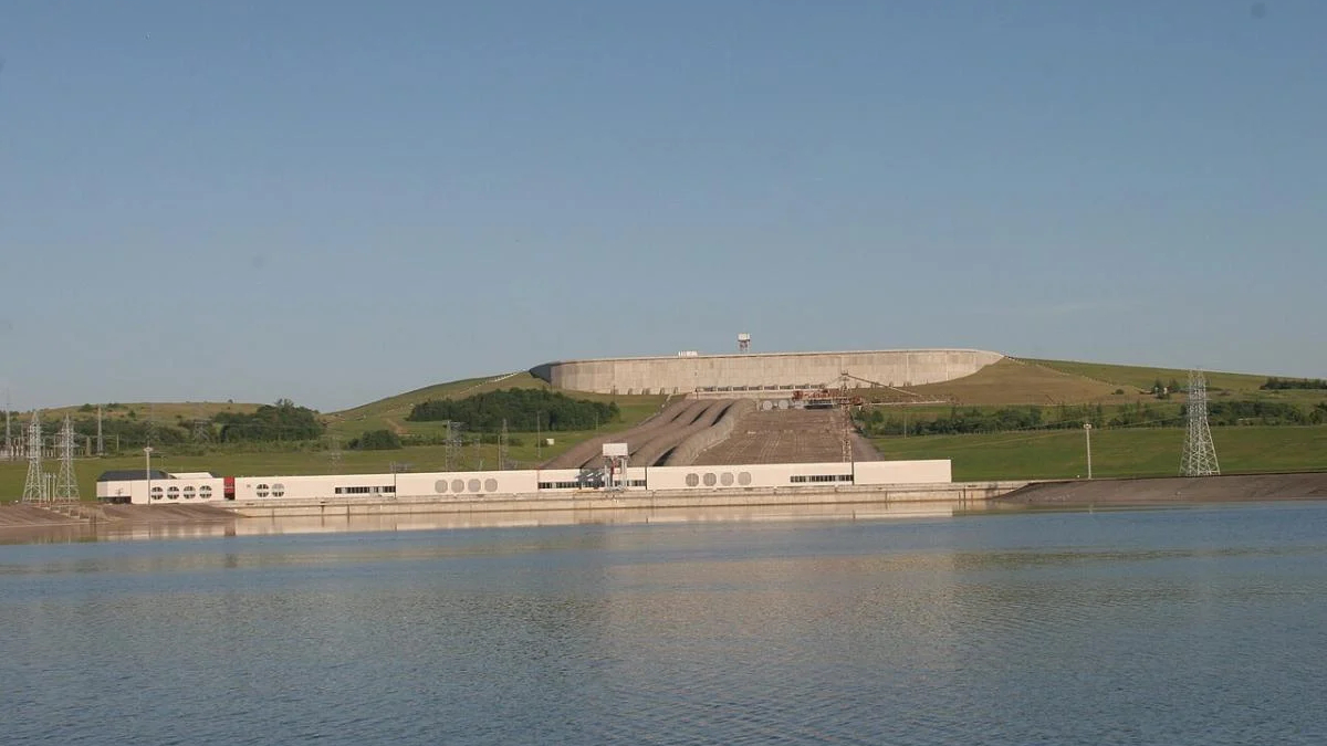 Hydroelectric facility by a lake with a long white building and grassy hill behind a concrete dam structure.