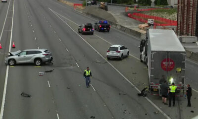 Highway crash scene with a silver SUV burned-out on the left, a white SUV in the middle, and a large box truck pulled over on the right as responders work nearby.