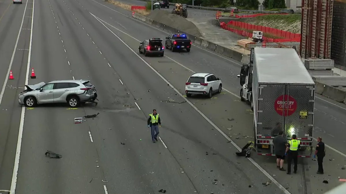 Highway crash scene with a silver SUV burned-out on the left, a white SUV in the middle, and a large box truck pulled over on the right as responders work nearby.