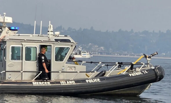 Police patrol boat on calm water with officer standing at the cabin door, blue emergency lights visible on top.