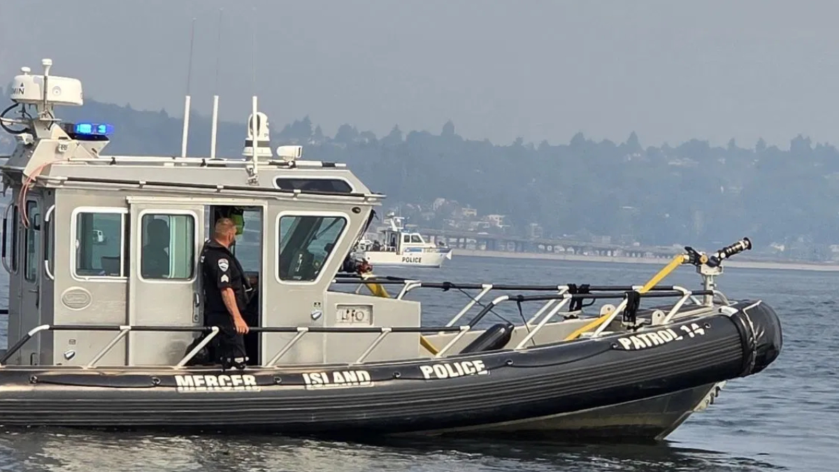 Police patrol boat on calm water with officer standing at the cabin door, blue emergency lights visible on top.