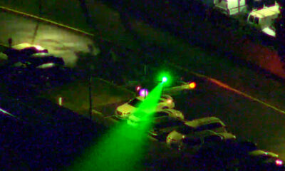 Nighttime street scene with a bright green beam or flashlight sweeping across parked cars toward a green-lit traffic signal.