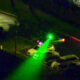 Nighttime street scene with a bright green beam or flashlight sweeping across parked cars toward a green-lit traffic signal.