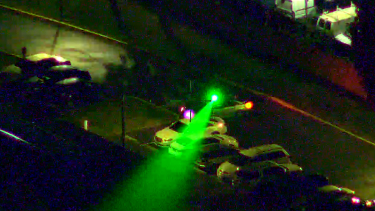 Nighttime street scene with a bright green beam or flashlight sweeping across parked cars toward a green-lit traffic signal.