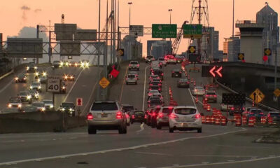 Cars in heavy traffic on a multi-lane bridge at sunset, with orange construction cones and signs in the roadway barrier area.