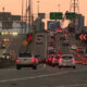 Cars in heavy traffic on a multi-lane bridge at sunset, with orange construction cones and signs in the roadway barrier area.