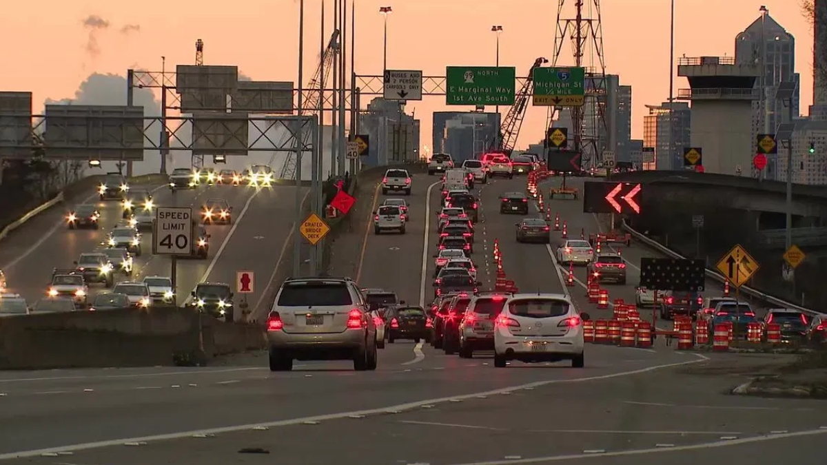 Cars in heavy traffic on a multi-lane bridge at sunset, with orange construction cones and signs in the roadway barrier area.