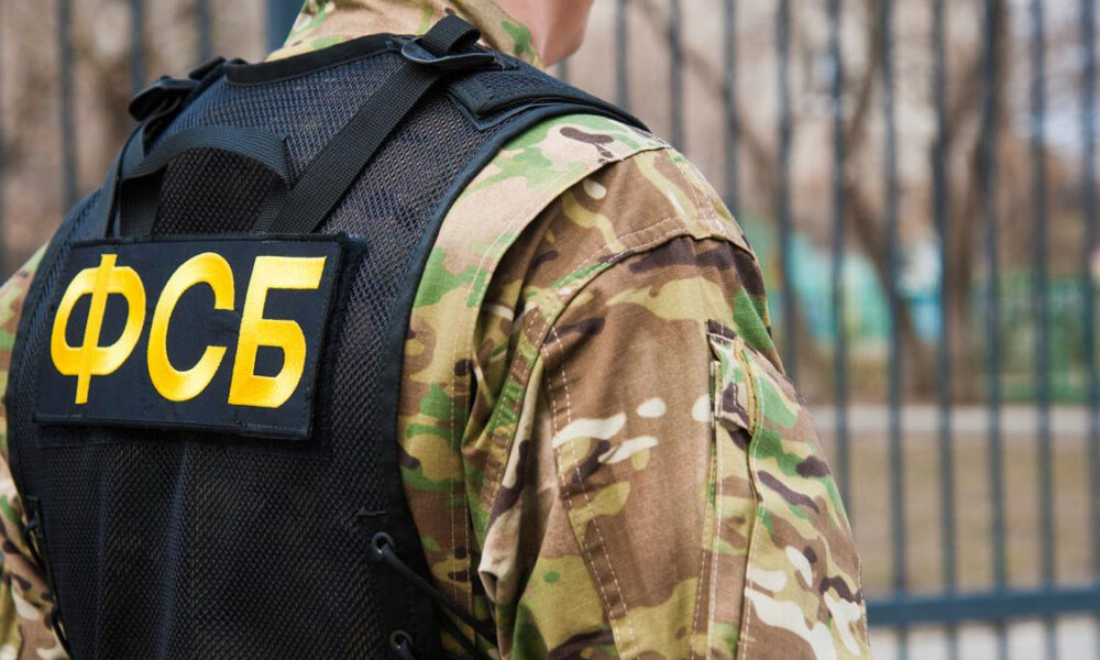Person in camouflage wearing a black tactical vest with a prominent yellow ФСБ patch on the back, outdoors near a fence