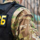 Person in camouflage wearing a black tactical vest with a prominent yellow ФСБ patch on the back, outdoors near a fence