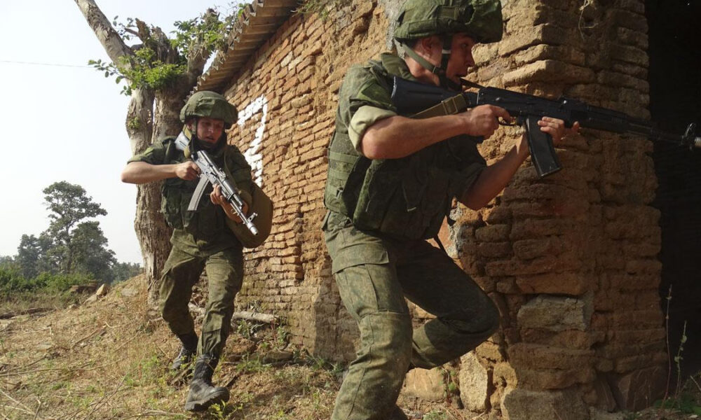 Two soldiers in olive uniforms advance with rifles beside a brick wall in a rural area.