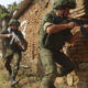 Two soldiers in olive uniforms advance with rifles beside a brick wall in a rural area.