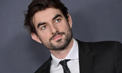 Professional headshot: man in a black suit and white shirt, looking to the left against a gray backdrop