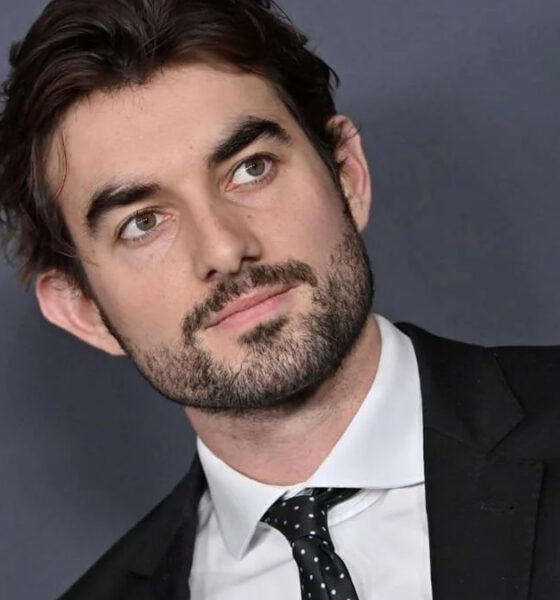 Professional headshot: man in a black suit and white shirt, looking to the left against a gray backdrop